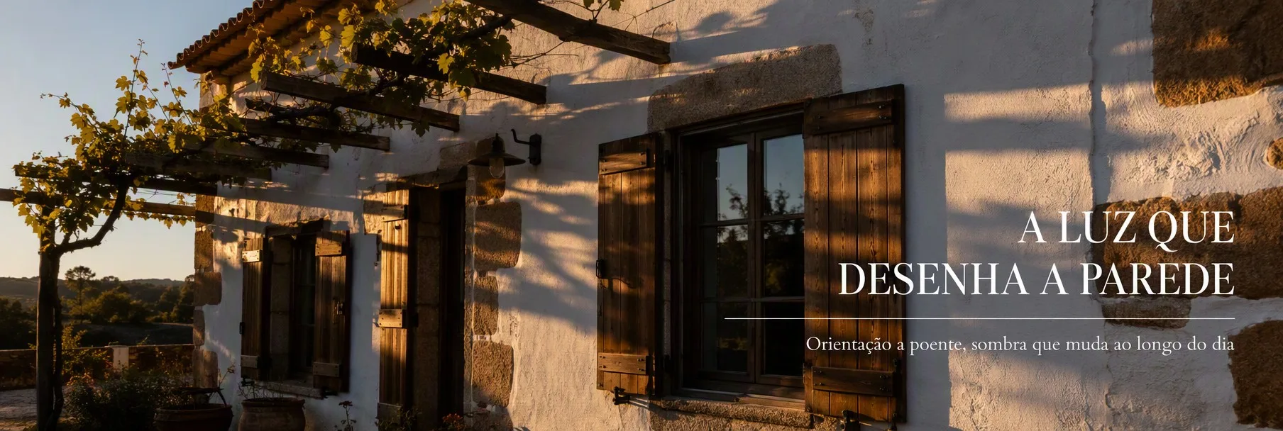 Fachada rústica portuguesa ao entardecer, com sombras de portadas e pérgola desenhadas na parede de cal